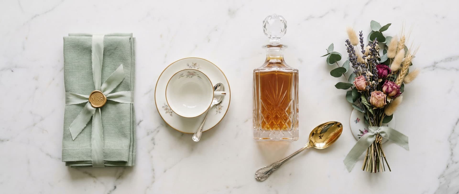 Flat lay de presentes de casamento com guardanapo sage, xicara de porcelana, licoreira de cristal e buque